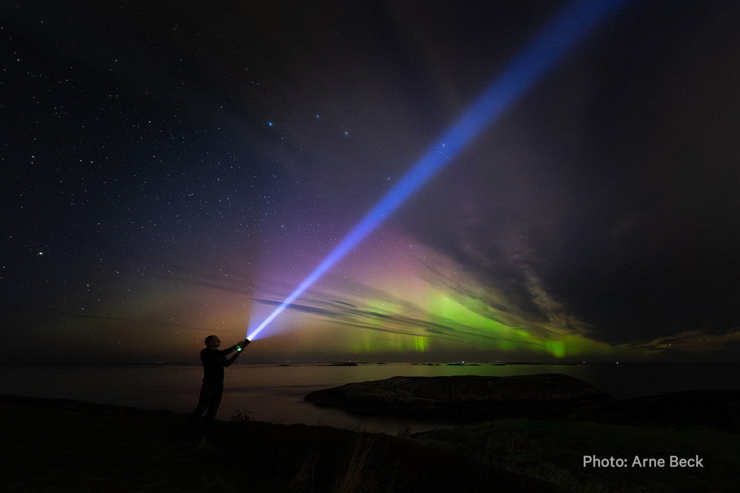 Person lyser mot kveldshimmelen under kraftig nordlys ved kysten. Foto: Arne Beck.