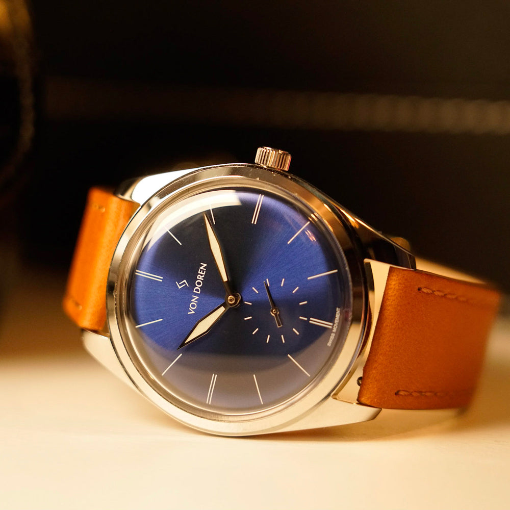 Von Doren-klokke med dyp blå sunburst-tallskive, sølvvisere og brun lærrem, fotografert i varm belysning.