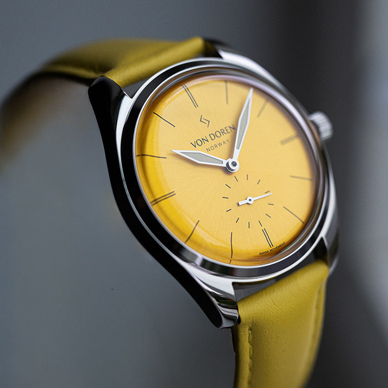 Von Doren klokke med gul sunburst-skive, gul skinnrem og polert stålkasse.