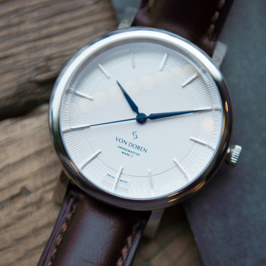 Von Doren klokke med hvit skive og blå visere med treverk i bakgrunnen.