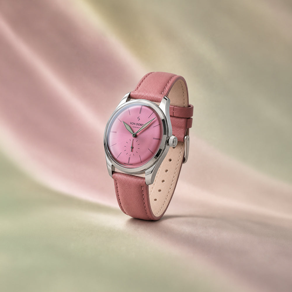 Von Doren-klokke med rosa skive, kasse i polert stål og rosa skinnrem med dus bakgrunn i rosa og grønn.