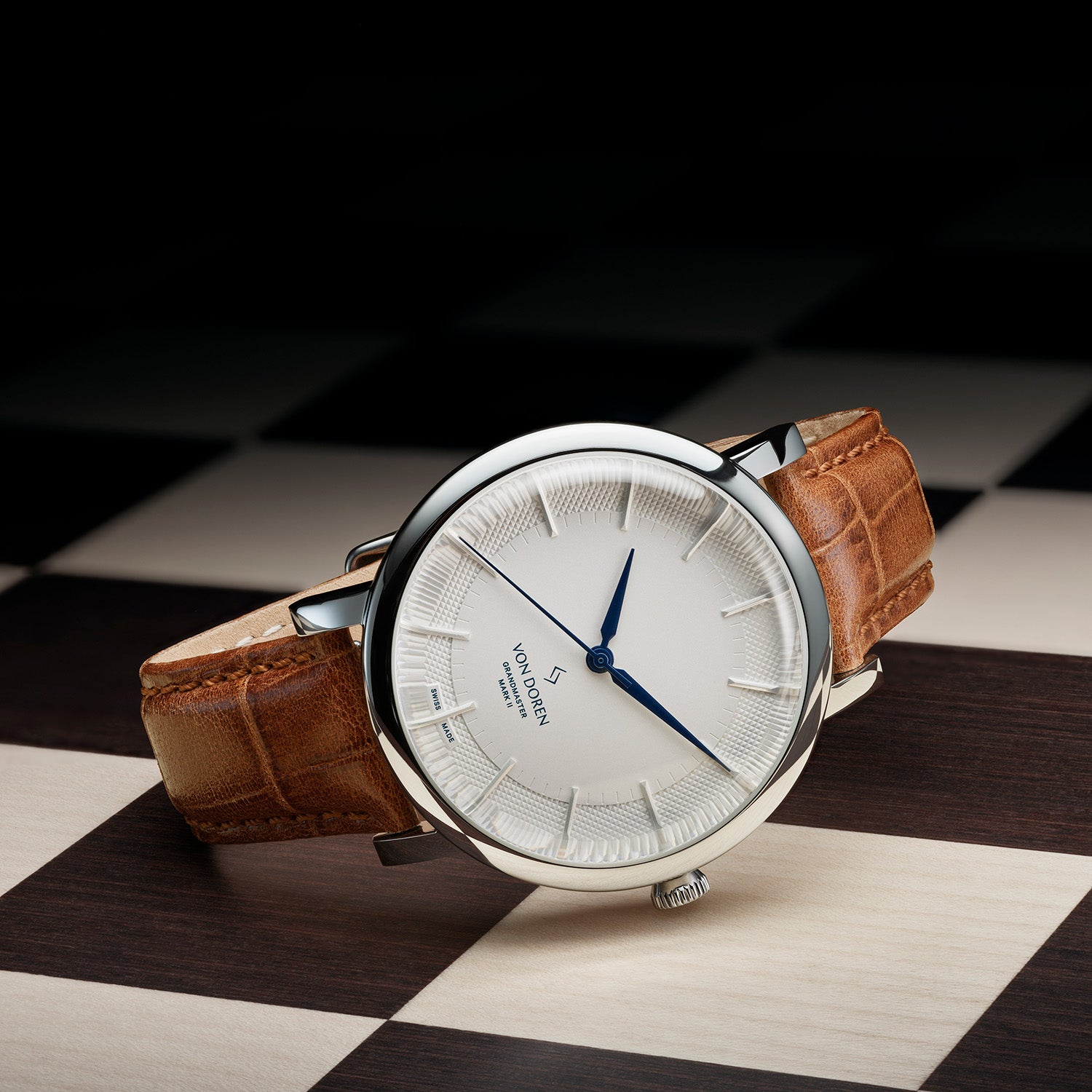 Von Doren-klokke med hvit tallskive og brun rem på sjakkmønstret bakgrunn.
